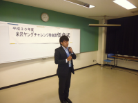 正面に「米沢ヤングチャレンジ特命課任命式」と書かれた紙が設置された会場で、1名の男性が挨拶をしている様子の写真