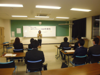 正面で1名の女性が挨拶をしているなか、椅子に着席した関係者たちが女性の方を向いて見守っている様子の写真