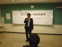 着席した関係者が見守る中、会場正面に立つ1名の男性が挨拶をしている様子の写真