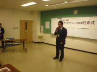 正面に「米沢ヤングチャレンジ特命課任命式」と書かれた紙が設置された会場で、スーツ姿の1名の男性が挨拶をしている様子の写真