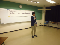 正面に「米沢ヤングチャレンジ特命課任命式」と書かれた紙が設置された会場で、ジャケット姿の1名の男性が挨拶をしている様子の写真