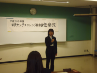 正面に「米沢ヤングチャレンジ特命課任命式」と書かれた紙が設置された会場で、1名の女性が挨拶をしている様子の写真