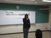 正面に「米沢ヤングチャレンジ特命課任命式」と書かれた紙が設置された会場で、1名の女性がマイクを手に挨拶をしている様子の写真
