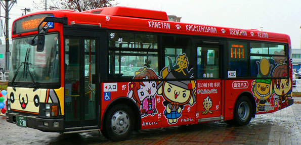 赤を基調とした、かねたん・おせんちゃん・かげっちさま・けーじろーがデザインされたラッピングバスの写真